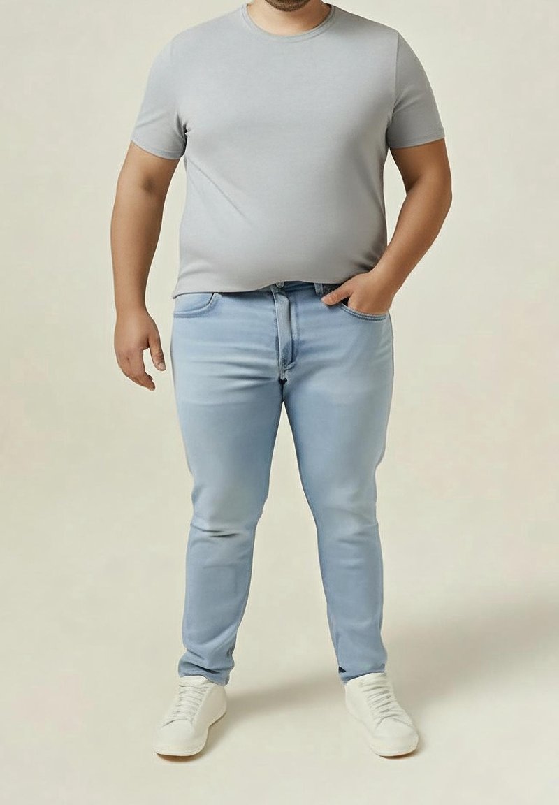 Homme portant un t-shirt gris, un jean bleu clair et des baskets blanches, debout avec une main dans sa poche.