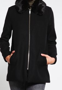 Abrigo negro con cuello de piel sintética, con cremallera frontal, bolsillos laterales y un corte entallado. Tela suave con una textura agradable.