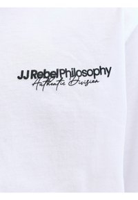 Λευκό ύφασμα με μαύρο κεντημένο κείμενο «JJ Rebel Philosophy» με έντονα γράμματα και «Authentic Division» σε πλάγια γραφή από κάτω.