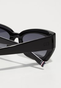 Black oversized sunglasses with angular frame, dark lenses, glossy finish, and distinct temple accents featuring a red, white, and blue stripe.