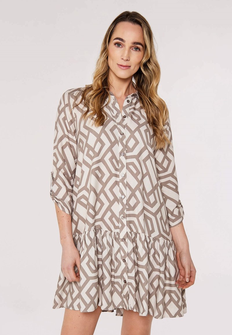 Frau mit langen welligen Haaren, die ein beiges und weißes, geometrisch gemustertes Kleid mit Knopfleiste und 3/4-Ärmeln trägt, steht vor einem schlichten Hintergrund.