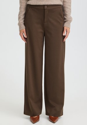Femme portant un pantalon marron taille haute à jambes larges avec des boutons à l'avant et des poches fendues, associé à des chaussures marron à bout pointu et un pull beige.
