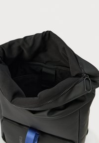 Μαύρο σακίδιο με ανοιχτό φερμουάρ στην κορυφή, εσωτερική επένδυση με επένδυση και μπροστινή τσέπη με φερμουάρ με μπλε υφασμάτινη ετικέτα τραβήγματος, παρουσιάζεται σε λευκό φόντο.