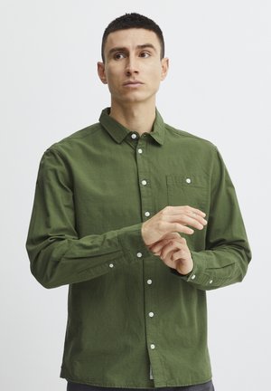 Camisa verde de botones hecha de una tela suave, con un bolsillo en el pecho, botones blancos y mangas largas con puños abotonados.