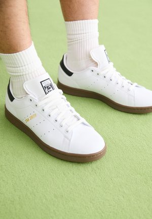 Osoba nosí bílé tenisky Adidas Stan Smith s černou patní částí a hnědou podrážkou, doplněné bílými žebrovanými ponožkami na zeleném povrchu.