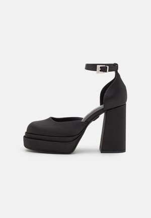 Zapato negro de tacón alto con plataforma, con un grueso tacón cuadrado y correa en el tobillo que presenta una hebilla plateada, mostrado de lado sobre un fondo blanco.