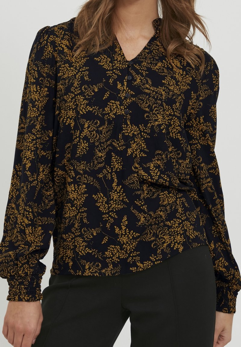 Femme portant un chemisier noir à manches longues avec un motif floral jaune moutarde et un pantalon noir.