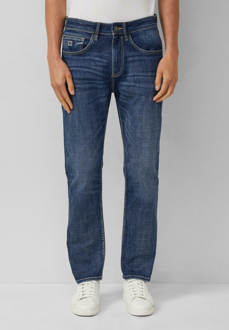 Dunkelblaue Denim-Jeans mit geradem Schnitt, genähten Details, Fünf-Taschen-Style und Metallknopfverschluss. Getragen mit weißen Sneakern.