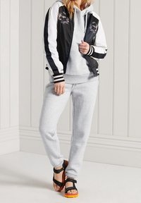 Giacca bomber bianca e nera con ricamo floreale, felpa grigia con cappuccio, pantaloni della tuta grigi e sandali neri con dettagli arancioni.