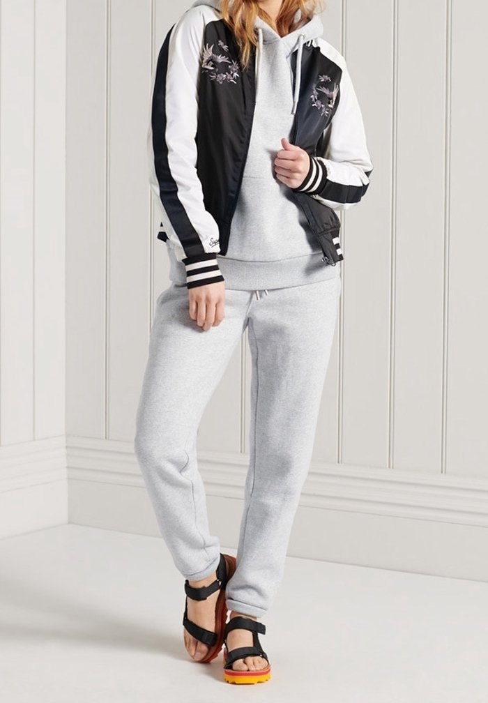 Giacca bomber bianca e nera con ricamo floreale, felpa grigia con cappuccio, pantaloni della tuta grigi e sandali neri con dettagli arancioni.