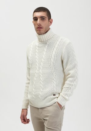 Suéter blanco crema de punto con un alto cuello acanalado y patrones texturizados, que presenta una mezcla de puntos de cable y puntos de palomitas.