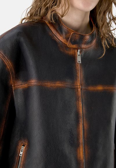 Veste en cuir noire avec des accents orange et des fermetures éclair, portée par une personne aux cheveux ondulés châtain clair, cadrée au niveau du cou et de la poitrine.