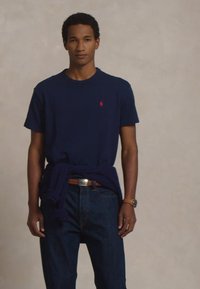 Fiatal férfi sötétkék pólót visel, piros logóval, sötét farmernadrággal, övvel, karórával, és egy derékra kötött pulóverrel egy sima háttér előtt.