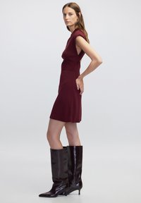 Vínová přiléhavá šaty s krátkými rukávy a bočními výřezy, doplněné vysokými lesklými černými koženými botami s špičatou špičkou a nízkým podpatkem.