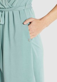 Vêtement bleu clair à coupe décontractée, doté d'une poche latérale et d'un petit logo argenté. Le tissu semble doux et fluide.