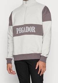 Sudadera gris claro con cuello alto, con una franja horizontal marrón y "PEGADOR" bordado en blanco. Puños y dobladillo acanalados.