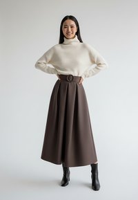 Kremowy prążkowany sweter z golfem oraz brązowa plisowana spódnica midi z okrągłym paskiem, zestawione z czarnymi botkami za kostkę.