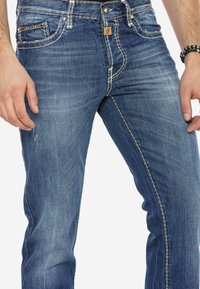 Denim jeans in middelblauw met subtiele vervaging, gouden stiksels, vijf zakken en een merklabel aan de voorkant. Veelzijdig en duurzaam ontwerp.