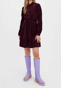 Abito camicia in corduroy color bordeaux con bottoni davanti, colletto e cintura abbinata. Indossato con stivali lavanda fino al ginocchio con suole testurizzate.