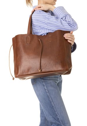 Vrouw in blauw gestreept overhemd en spijkerbroek die een grote bruine leren handtas over haar schouder draagt.