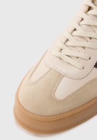 Beige sneaker with leather and suede upper; features cream laces, soft texture, and a gum sole. Accent stitching and decorative edges visible.