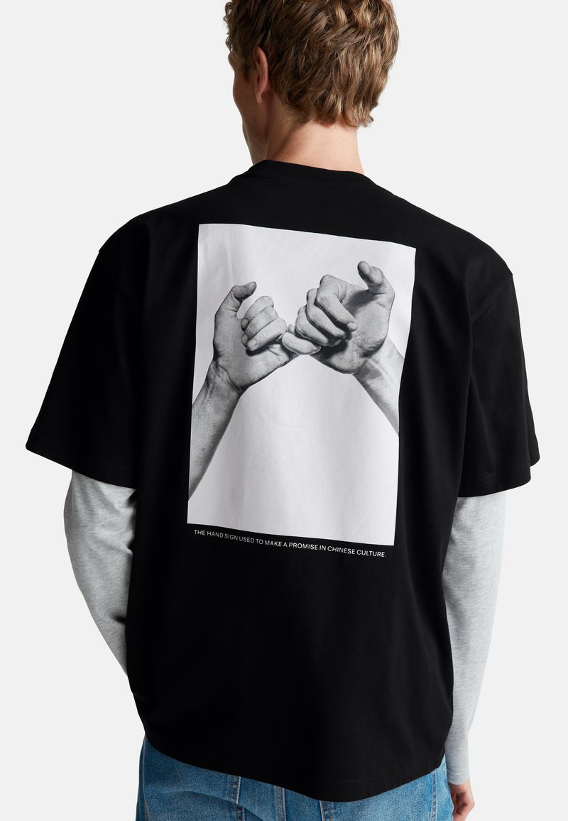Muž v černém tričku s černobílým potiskem dvou rukou, které si dělají malíčkový slib, stojící na jednolitém pozadí.