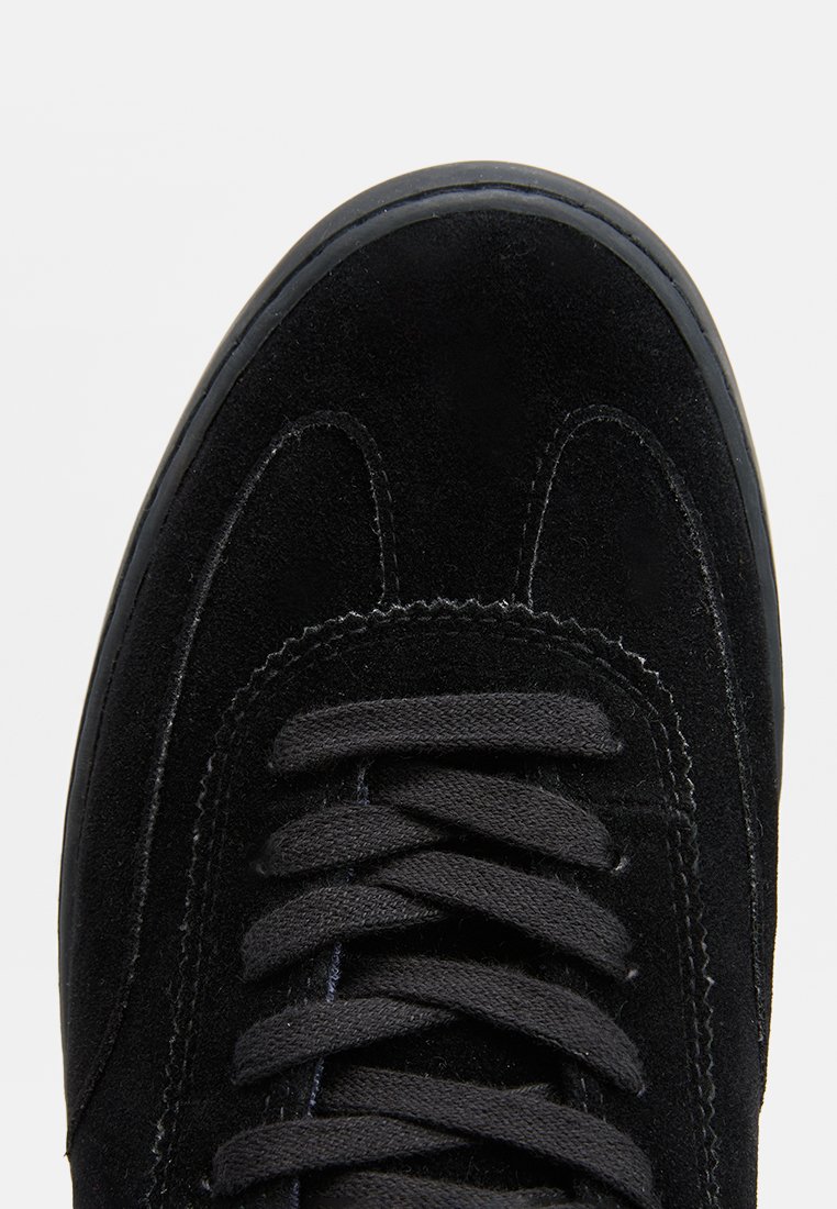 Vista superior de una sneaker de ante negra, con punta redondeada, cordones negros y una superficie texturizada con sutiles detalles de costura.