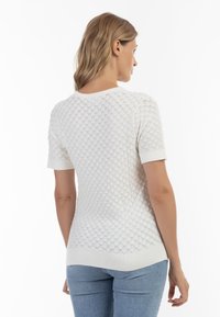 Hvid strikket sweater med et struktureret skælmønster, korte ærmer og en rund halsudskæring. Bæres med blå tætsiddende jeans.