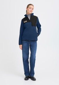 Pullover in pile blu con pannello frontale con zip nera, colletto alto e dettaglio a patch. Indossato con jeans blu e calzature nere.