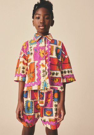 Kind trägt ein bunt gemustertes Hemd- und Shorts-Set mit Obst- und Blumenmotiven, steht vor einem schlichten beige-farbenen Hintergrund.