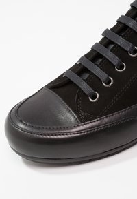 Section avant d'une basket en cuir et daim noir avec des lacets noirs passés dans des œillets métalliques sur fond blanc.