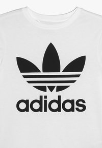 Balts kokvilnas T-krekls ar lielu melnu Adidas trefoil logo un zīmola nosaukumu bolds melnās burtos zem tā.