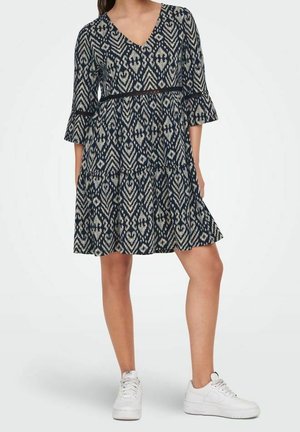 Femme portant une robe noire et blanche à motifs, longueur genou, avec des manches évasées trois-quarts, et des baskets blanches, debout devant un fond uni.