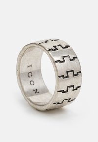 Anel de metal prateado com um padrão geométrico em zigzag gravado ao redor da banda, com uma textura suave e um acabamento mate.