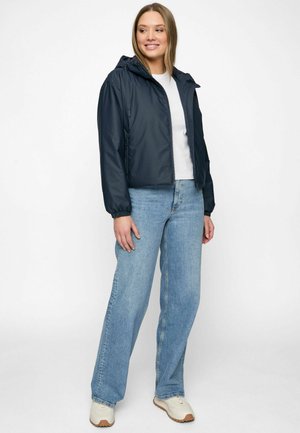 Frau lächelnd, trägt eine schwarze Kapuzenjacke, weißes Oberteil, hellblaue Jeans mit weitem Bein und beige Turnschuhe, steht vor einem schlichten weißen Hintergrund.