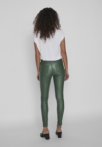 Pantalones de cuero sintético verdes, ajustados y de cintura alta, con bolsillos traseros y una textura brillante, combinados con una blusa blanca de manga corta y mules negros.