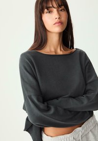 Graues Langarmoberteil mit weitem Ausschnitt und geripptem Design. Lockere Passform mit Abnähern an den Schultern. Kombiniert mit hellgrauen, lässigen Hosen.
