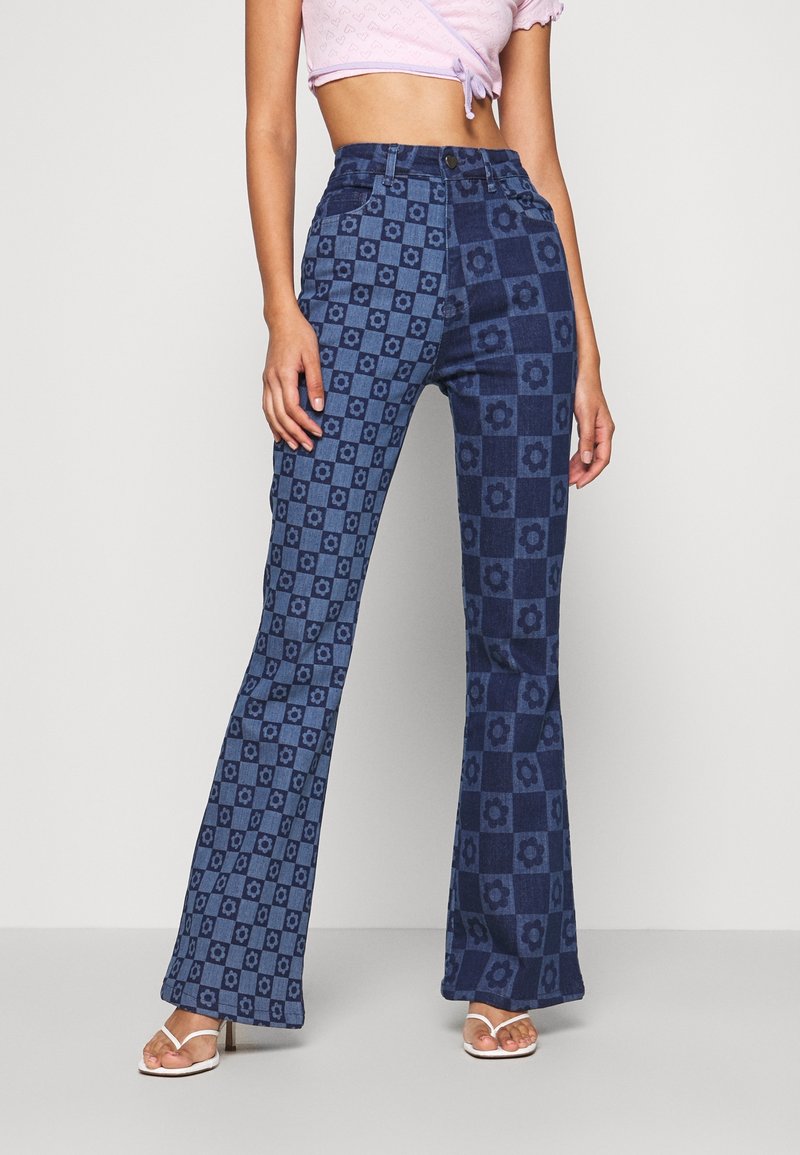 Femme portant un jean flare taille haute avec imprimé floral à carreaux bleu clair et bleu foncé et des sandales blanches à talons.