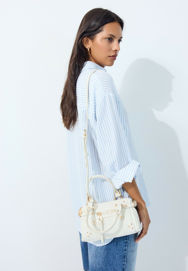 Femme aux longs cheveux foncés portant une chemise à rayures bleues et un jean, avec un petit sac à main blanc à ferrures dorées sur l'épaule.