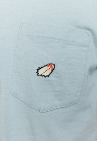 Lichtblauw katoenen t-shirt met een geborduurd wit en rood raketembleem op de linkerzak. Zachte textuur, standaard ronde hals, korte mouwen.