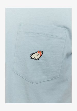 Light blue cotton t-shirt featuring a stitched white and red rocket emblem on the left pocket. Soft texture, standard crew neck, short sleeves.