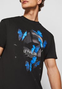 Sort kortærmet T-shirt med et grafisk design af blå sommerfugle og teksten "LE PAPILLON" i hvid. Blødt bomuldsstof.