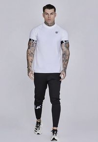 Hvid kortærmet T-shirt med sort logo og stribede detaljer, kombineret med sorte tætsiddende joggers med hvid tekst på siden. Sneakers fuldender looket.