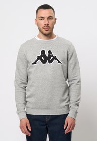 Világosszürke, pamut keverék pulóver, elöl fekete grafikai mintával, amely két ülő alakot ábrázol. Kerek nyakkivágás és bordázott mandzsetták jellemzik.