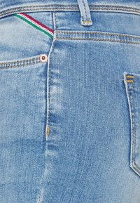 Jeans de ganga azul claro com uma superfície texturizada, apresentando costuras contrastantes, um pequeno bolso com riscas vermelha, branca e verde, e um rebite metálico.