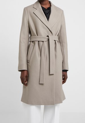 Beżowy wełniany płaszcz z paskiem, kołnierzem z klapami, noszony na czarnej bluzce i białych spodniach, z frontowymi i bocznymi kieszeniami, długimi rękawami.