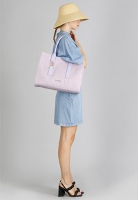 Borsa a tracolla lilla con tessuto testurizzato, doppi manici superiori e un accento dorato. Indossata su un vestito di denim, abbinata a un cappello a tesa larga.