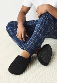 Papuci negri pufoși, cu o formă rotundă și o textură moale. Talpa are un model geometric și sunt purtați cu pantaloni de pijama în carouri bleumarin.