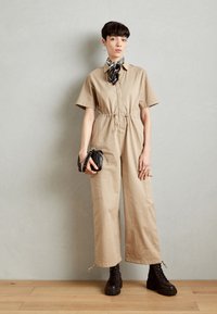 Beige cotton jumpsuit with short sleeves, cinched waist, and large pockets. Paired with black lace-up boots and a patterned scarf.