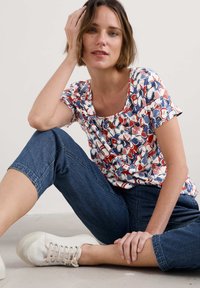 Tričko s krátkým rukávem a kulatým výstřihem s květinovým vzorem v červené, námořnické a bílé barvě. Spárováno s tmavě modrými zkrácenými kalhotami a bílými teniskami.
